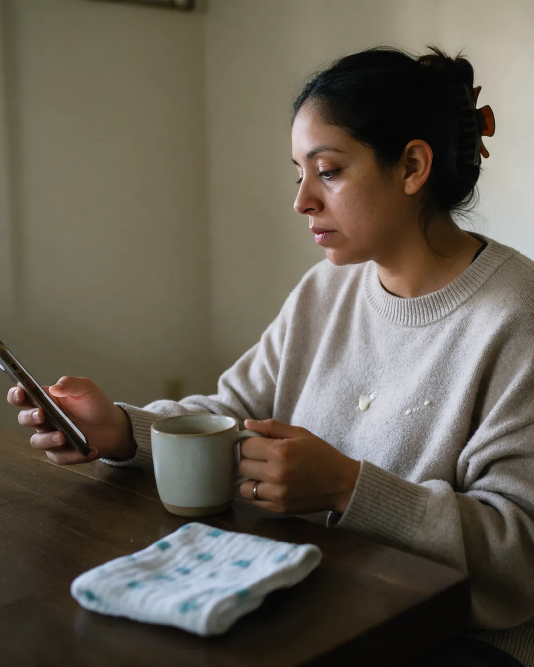 A new mother checks her phone at the kitchen table while her baby sleeps beside her.