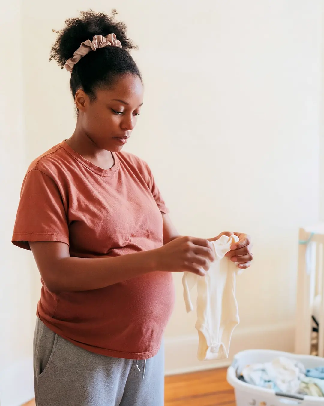 A pregnant woman folding baby clothes in a sunlit nursery